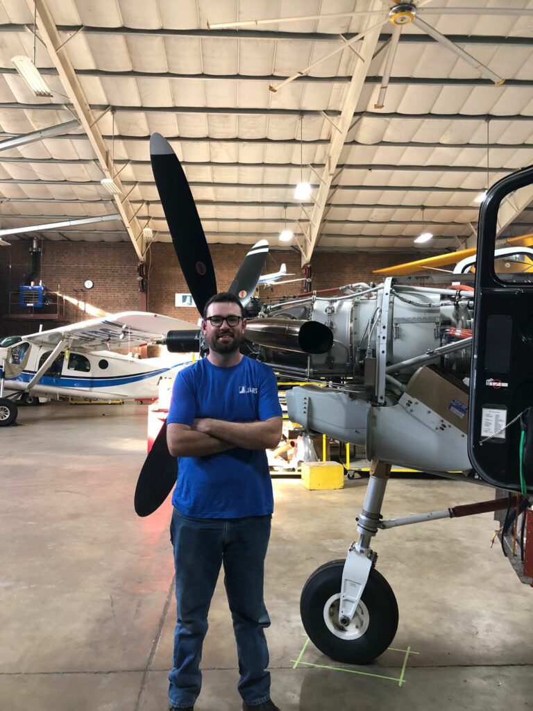 A white man in his thirties stands in front of an exposed Kodiak engine in an airplane hangar.