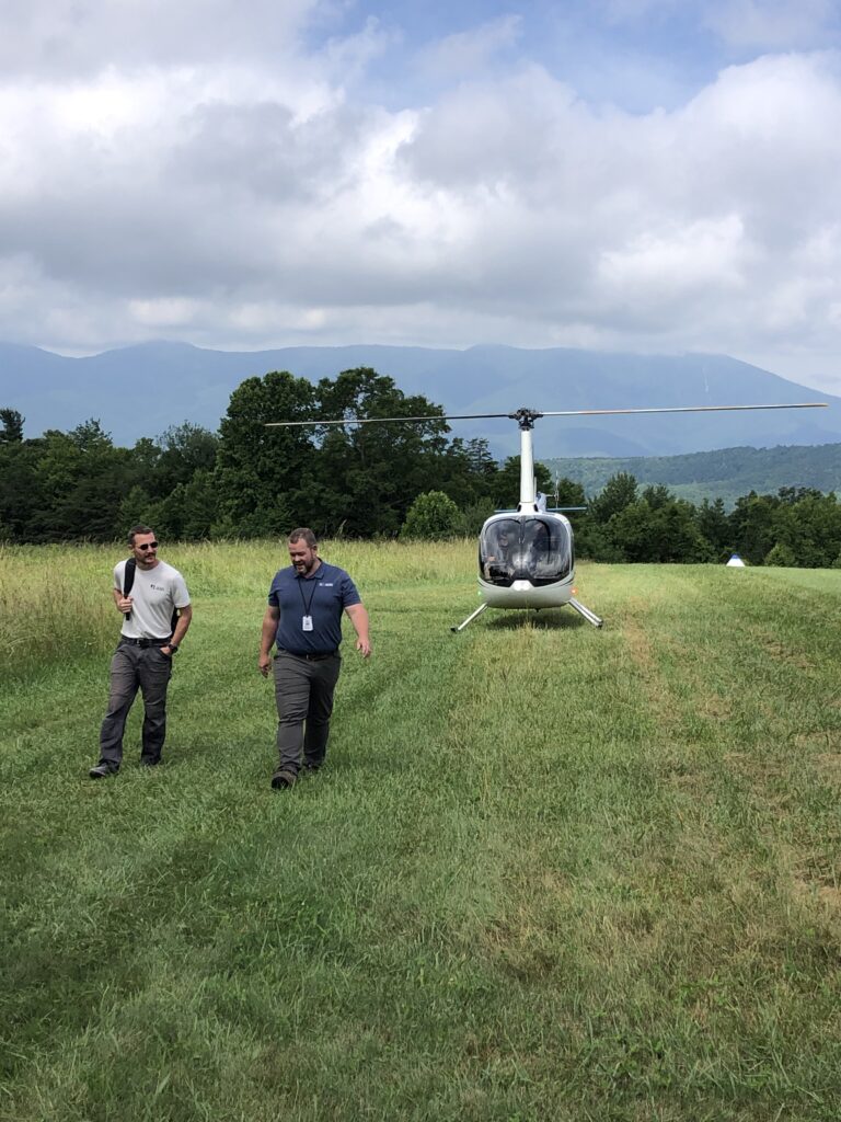 Two JAARS pilots walk in front of an R66 helicopter on top of a grass runway with the North Carolina Mountains behind them.