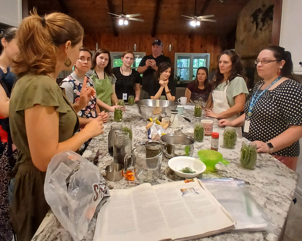 A group of women listen as another women tells them how to pickle green beans around a kitchen counter.