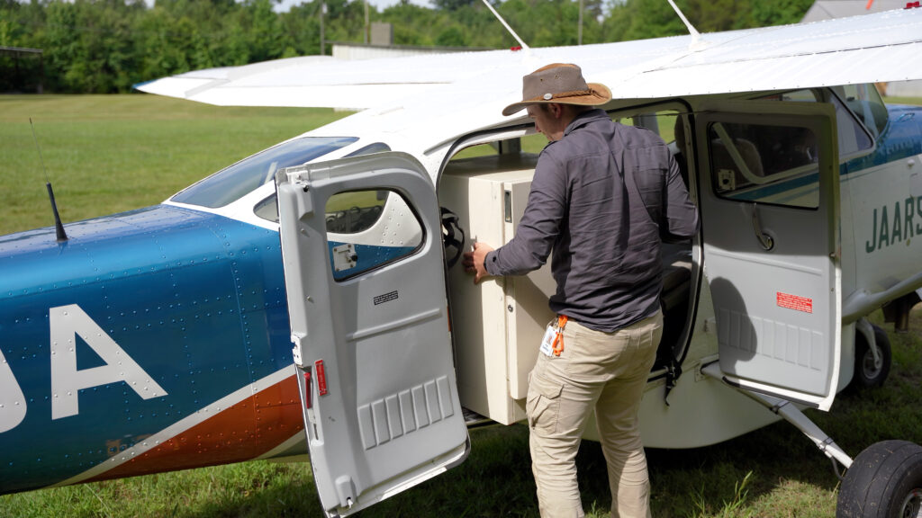 A pilot loads a filing cabinet into the back of a Cessna 206 outside.